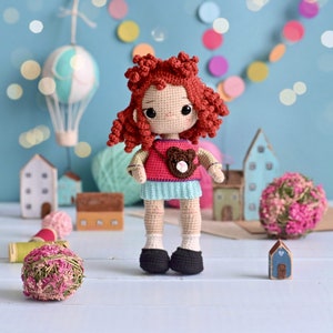 May include: A crocheted doll with red hair, wearing a pink and blue dress and a brown bear backpack. The doll is standing on a white surface with a blue background.