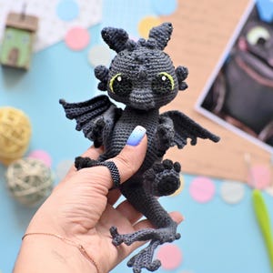 May include: A black crocheted dragon with green eyes and a light blue background. The dragon is being held by a hand with blue nail polish.
