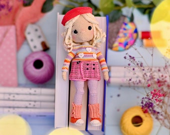 May include: A crocheted doll with blonde hair, wearing a pink and white striped dress, a red hat, and pink shoes. The doll is sitting on a stack of books.