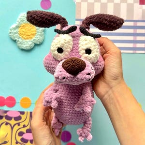 May include: A crochet plush toy of a cartoon dog with light purple fur, large white eyes, a dark brown nose, and dark brown floppy ears. The character has a worried expression with thick black eyebrows and small limbs, crafted from soft yarn.
