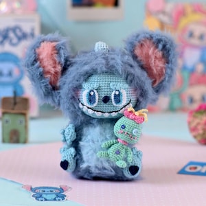 Może przedstawiać: Ręcznie robiona niebieska pluszowa zabawka na szydełku, przypominająca postać z kreskówki, z dużymi uszami i puszystą fakturą, trzymająca mniejszą zieloną szydełkową figurkę. Zabawka ma uśmiechniętą twarz i jest na różowo-niebieskim tle.