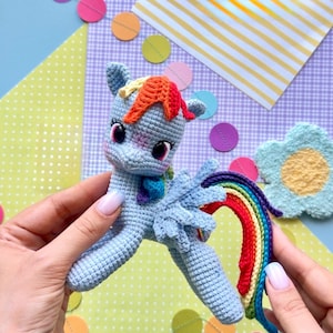 May include: A light blue crocheted pony toy featuring a vibrant rainbow-colored mane and tail, large dark eyes with long lashes, and small wings. The pony has a soft, textured yarn construction with detailed stitching throughout its body.