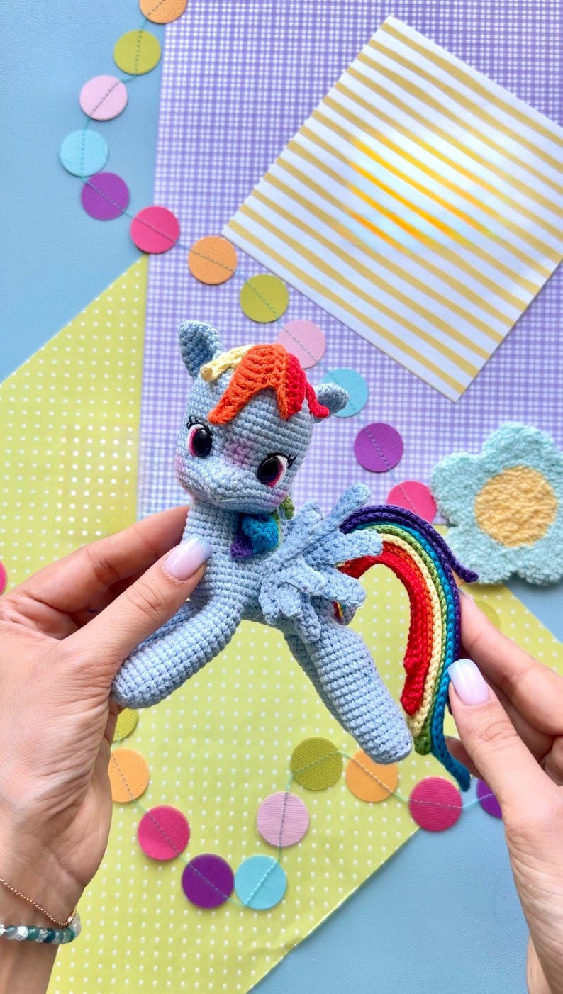 May include: A light blue crocheted pony toy featuring a vibrant rainbow-colored mane and tail, large dark eyes with long lashes, and small wings. The pony has a soft, textured yarn construction with detailed stitching throughout its body.