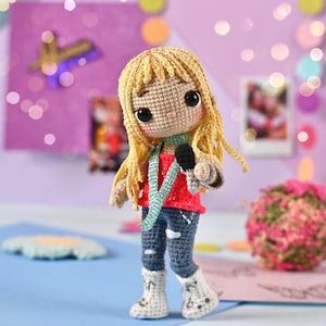 May include: A handmade crochet doll of a singer with blonde hair, wearing a red shirt, blue jeans, and white boots. The doll is holding a black microphone and has a green scarf. The background includes a purple board and a decorative ball.