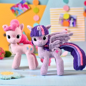 May include: Two crocheted pony figures. One is light pink with a darker pink mane and tail, featuring a blue and yellow cutie mark. The other is light purple with a dark purple mane, pink and blue highlights, wings, and a pink star cutie mark.