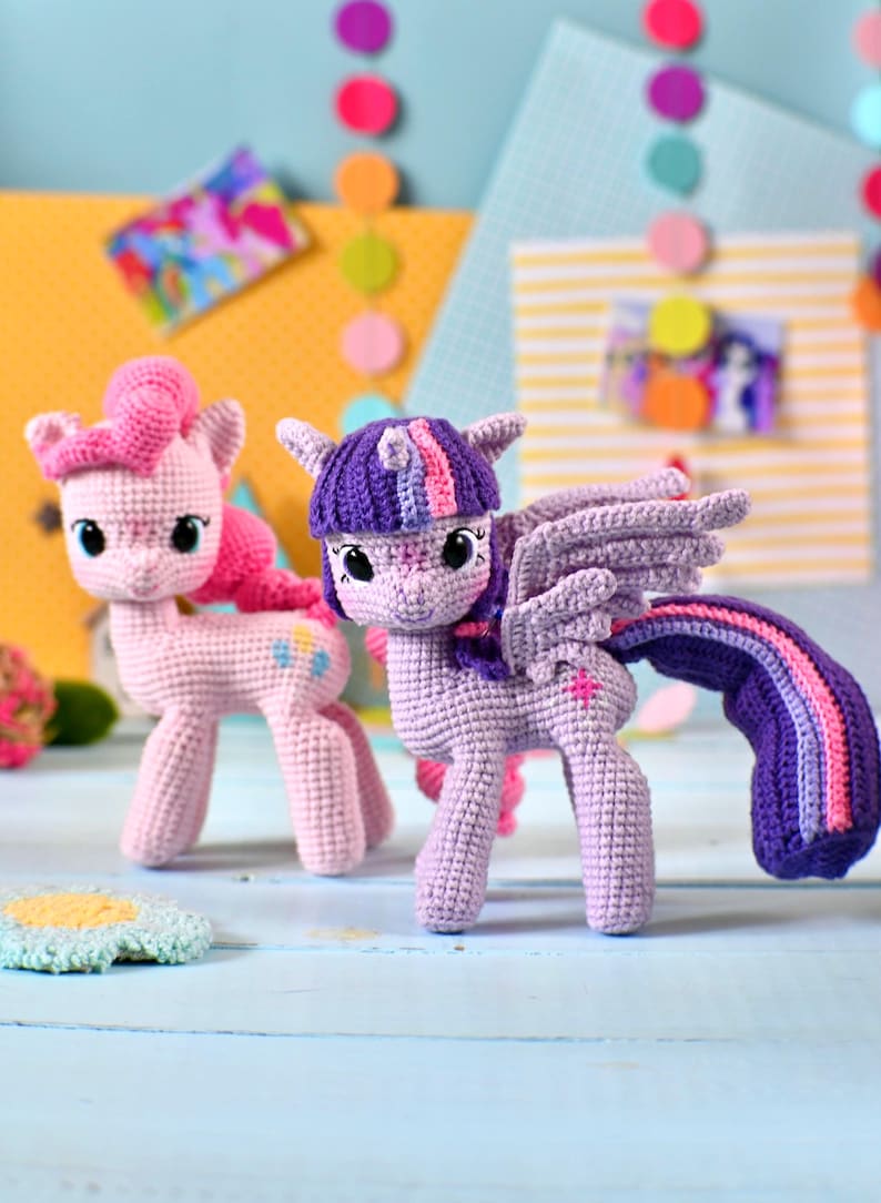 May include: Two crocheted pony figures. One is light pink with a darker pink mane and tail, featuring a blue and yellow cutie mark. The other is light purple with a dark purple mane, pink and blue highlights, wings, and a pink star cutie mark.