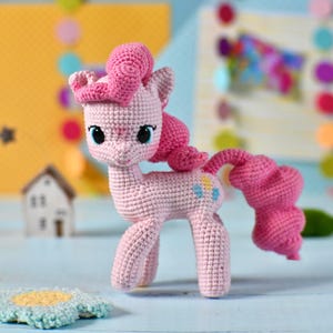 May include: A light pink crocheted pony figurine with a darker pink curly mane and tail. The pony features large black eyes with blue highlights and a small embroidered cutie mark on its flank consisting of two blue balloons and one yellow balloon.