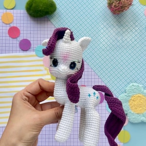 May include: A white crocheted unicorn toy with a dark purple mane and tail. The unicorn features large blue eyes, a small horn on its forehead, and three small blue star-shaped embroidered details on its flank. The yarn texture is visible.