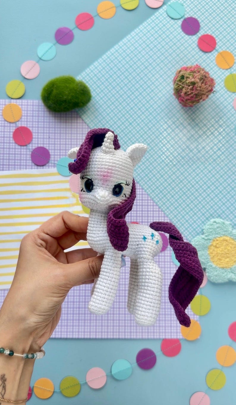 May include: A white crocheted unicorn toy with a dark purple mane and tail. The unicorn features large blue eyes, a small horn on its forehead, and three small blue star-shaped embroidered details on its flank. The yarn texture is visible.