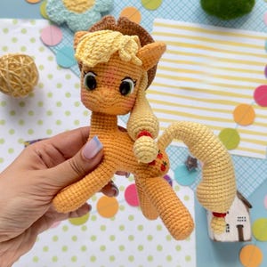 May include: A crocheted orange pony figurine featuring a light yellow mane and tail, a brown cowboy hat, and three small red apple icons embroidered on its flank. The toy has large dark eyes and a textured yarn construction throughout its body.