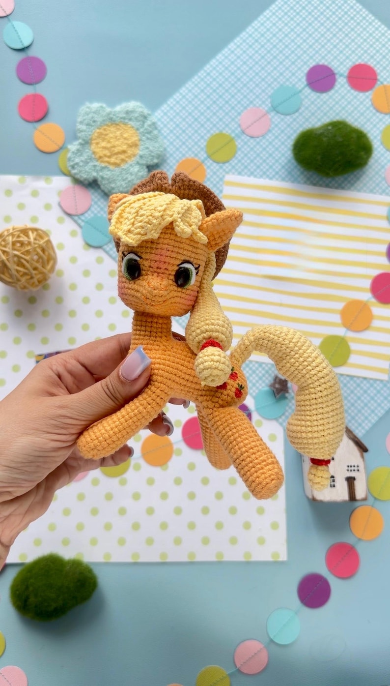 May include: A crocheted orange pony figurine featuring a light yellow mane and tail, a brown cowboy hat, and three small red apple icons embroidered on its flank. The toy has large dark eyes and a textured yarn construction throughout its body.