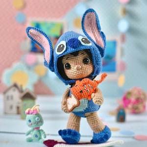 May include: A handmade crochet doll of a character wearing a blue costume with large ears, holding an orange object, and standing on a surfboard. A small green doll is nearby. The background is light blue and pink.