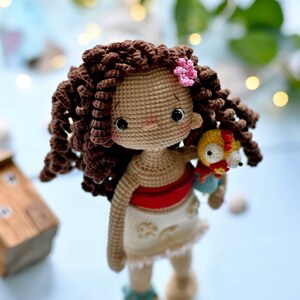 Op de afbeelding: Een gehaakte pop met bruin krullend haar, een rood topje, een witte rok en een klein gehaakt vogeltje in haar hand. De pop draagt een roze bloemetje in haar haar.