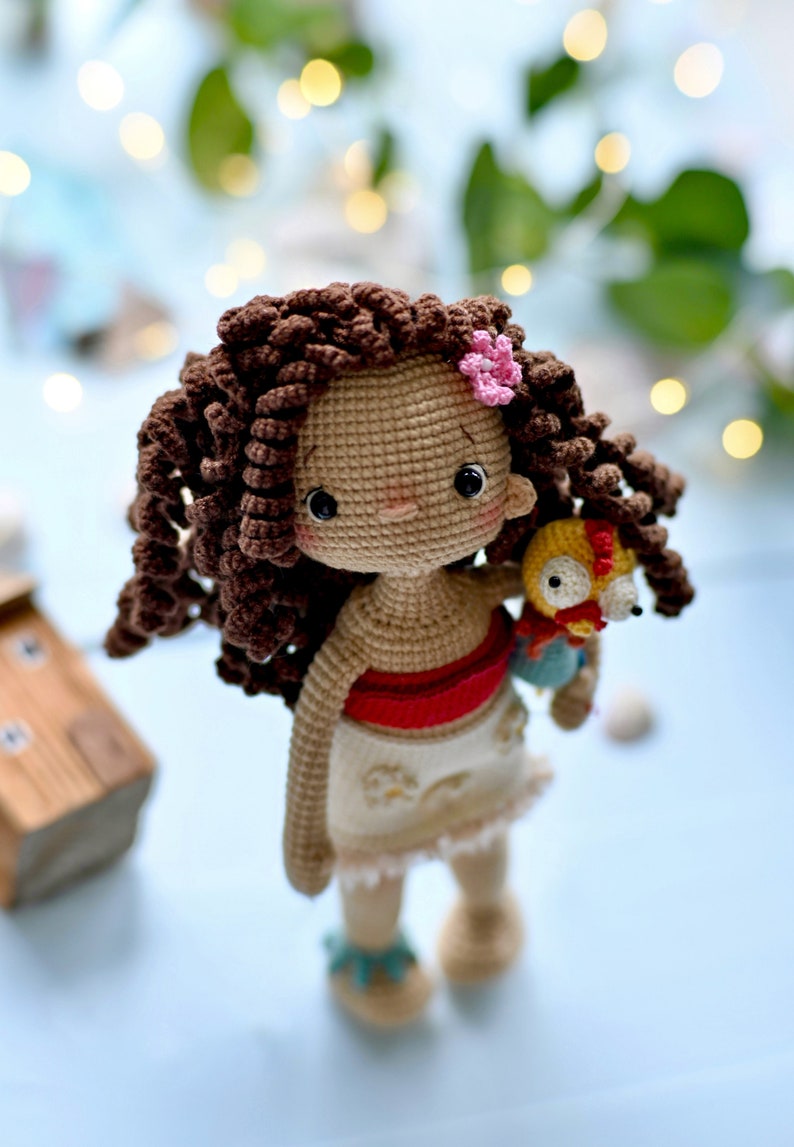 Op de afbeelding: Een gehaakte pop met bruin krullend haar, een rood topje, een witte rok en een klein gehaakt vogeltje in haar hand. De pop draagt een roze bloemetje in haar haar.