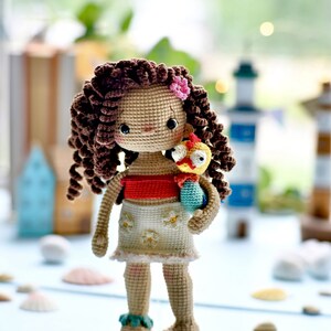 Op de afbeelding: Een gehaakte pop met bruin krullend haar, die een rood topje en een witte rok met bloemenmotief draagt, en een gehaakte gele en blauwe vogel vasthoudt.