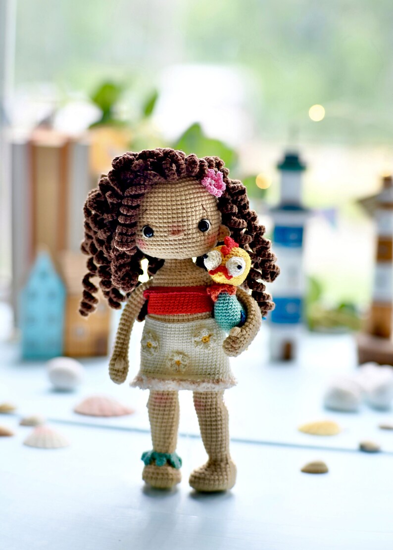Op de afbeelding: Een gehaakte pop met bruin krullend haar, die een rood topje en een witte rok met bloemenmotief draagt, en een gehaakte gele en blauwe vogel vasthoudt.