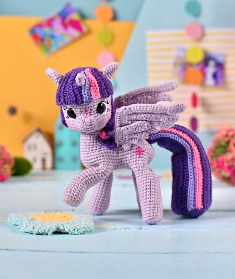 May include: A crocheted lavender pony figurine with large dark eyes, a purple mane featuring pink and blue stripes, and matching wings. The pony has a long, flowing purple tail with pink and blue stripes and a small pink star emblem on its flank.