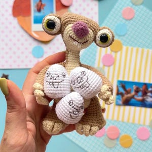 以下が含まれることがあります： 手作りの、映画「アイス・エイジ」のナマケモノ、シドのかぎ針編み人形。顔が描かれた白い卵を3つ持っています。人形は茶色で、紫色の鼻と大きな目があります。背景にはカラフルな紙の切り抜きがあります。