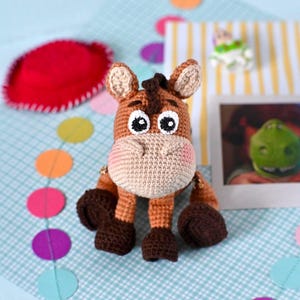 Puede incluir: Un juguete de burro hecho a mano con ganchillo, en tonos marrón, beige y negro. El burro tiene ojos grandes y expresivos, mejillas rosadas y una melena detallada. Está sobre una superficie a cuadros azul y blanco, con confeti colorido y un sombrero rojo.