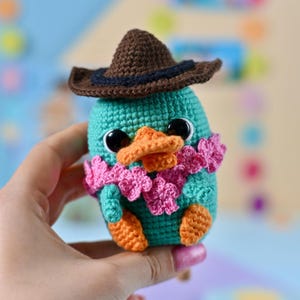Puede incluir: Un pato de ganchillo turquesa con pico y patas naranjas, que lleva un sombrero de vaquero marrón y una bufanda floral rosa. El pato tiene grandes ojos negros. El juguete hecho a mano se sostiene en una mano.