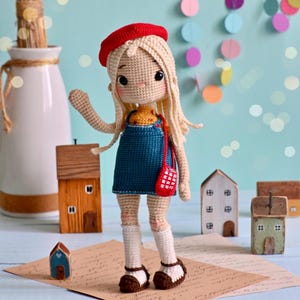 May include: A handmade crochet doll with blonde hair, a red beret, and a blue dress. The doll is wearing white socks and brown shoes. A small red purse hangs from its shoulder. Miniature wooden houses are in the background.