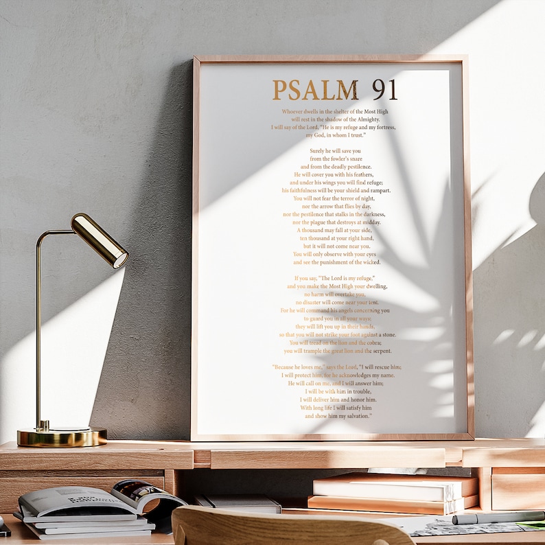 May include: A framed print of Psalm 91 in gold lettering. The text is displayed on a white background within a light brown frame. A gold desk lamp is on a wooden desk in the foreground.