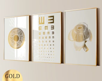 May include: Three gold framed prints with a white background. The left print features a stylized eye with gold detailing. The center print features a gold eye chart with the letters "E" and "W". The right print features a gold botanical illustration of a flower.