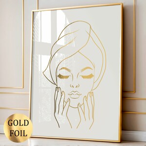May include: A gold foil line art print of a woman with her eyes closed and hands on her face. The woman is wearing a towel on her head.