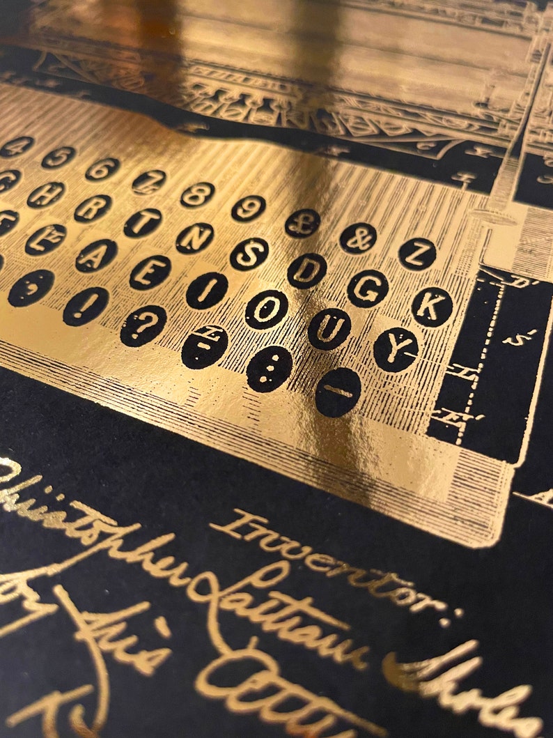 Peut inclure: Gros plan sur un clavier de machine &agrave; &eacute;crire couleur or avec les touches &eacute;tiquet&eacute;es avec des lettres et des chiffres. L'inscription "Inventor:  Latham Sholes" est visible en bas de l'image.