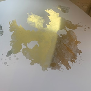May include: A white background with a large gold splatter of paint. The splatter has a rough, textured look and is slightly translucent.