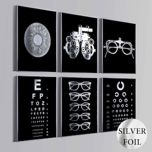 May include: Set of six black and silver foil framed art prints. The prints feature eye-related imagery, including an eye diagram, an eye exam machine, eyeglasses, an eye chart, and a vision test chart. The prints are labeled "Silver Foil".