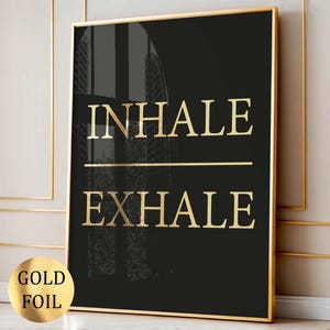May include: A rectangular wall art print featuring a solid black background with the gold foil text INHALE and EXHALE separated by a horizontal gold line. The print is encased in a thin gold frame and includes a circular gold foil label.