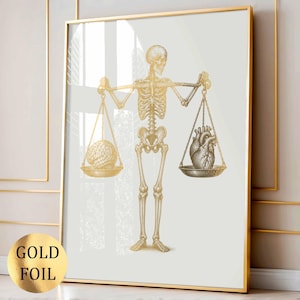 Könnte beinhalten: Ein gerahmter Wandkunstdruck mit einer Goldfolienillustration eines menschlichen Skeletts, das eine Waage hält. Auf einer Seite der Waage liegt ein menschliches Gehirn, auf der anderen Seite ein menschliches Herz. Der Hintergrund ist cremeweiß.