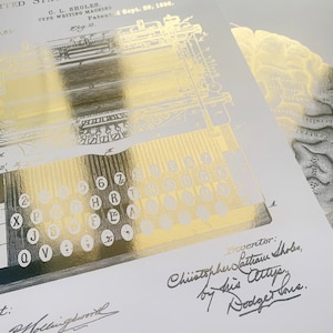 May include: A gold foil print on white paper featuring a patent illustration of a C. L. Sholes type writing machine from 1896. The design includes technical diagrams, a keyboard layout, and the handwritten signature of inventor Christopher Latham Sholes.