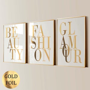 Puede incluir: Tres impresiones artísticas enmarcadas con letras doradas en lámina de oro que deletrean "BEAUTY", "FASHION" y "GLAMOUR" sobre un fondo blanco. Los marcos tienen un fino borde dorado. Una pegatina de lámina dorada está en la esquina inferior izquierda.