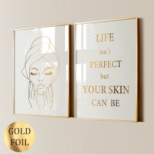 May include: Two framed prints with gold foil lettering. The left print features a line drawing of a woman with her eyes closed, touching her face. The right print has the text "LIFE isn't PERFECT but YOUR SKIN CAN BE".