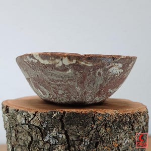 May include: A handcrafted, marbled ceramic bowl with a reddish-brown and cream color scheme. The bowl has a unique, organic shape and sits on a rustic wooden base, showcasing its natural texture and design.
