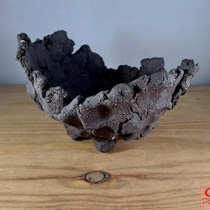 Pode incluir: Um vaso de cerâmica preto com uma superfície áspera e texturizada. O vaso tem uma forma irregular, assimétrica e uma abertura larga e rasa.