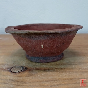 Könnte beinhalten: Eine kleine, runde Keramikschale in Terrakotta-Farbe mit einem breiten, ausgestellten Rand und einer texturierten, matten Oberfläche. Die Schale hat einen dunklen, verwitterten Boden und ein leicht unebenes, handgefertigtes Aussehen in Rotbraun.