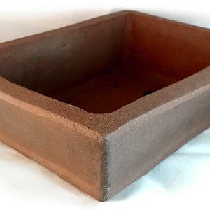 Könnte beinhalten: Ein rechteckiger brauner Keramik-Bonsai-Topf mit rauer Textur. Der Topf hat einen breiten, flachen Boden und einen leicht gekrümmten Rand.