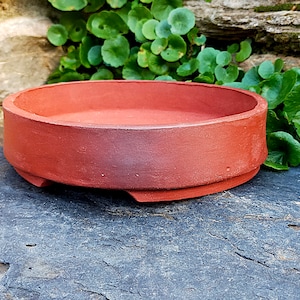 Könnte beinhalten: Ein runder Bonsai-Topf aus Terrakotta mit leicht ausgestelltem Rand und flachem Boden. Der Topf ist unglasiert und hat eine rotbraune Farbe.