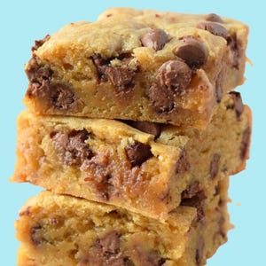 May include: A stack of four chocolate chip cookie bars with a light brown color and chocolate chips throughout. The bars are stacked on a light blue background.