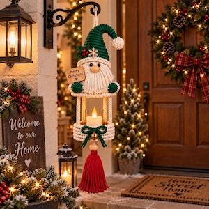 Könnte beinhalten: Eine Makramee-Gnom-Laterne mit einer grünen Mütze, einem cremefarbenen Bart und einem Holzschild mit der Aufschrift Merry Christmas. Die Struktur umfasst einen zentralen Glaskerzenhalter, eine grüne Schleife mit Goldglocke und eine rote Quaste.