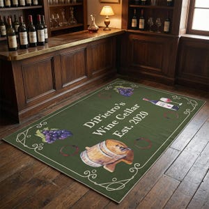 Op de afbeelding: Een rechthoekig groen tapijt met de tekst "DiPietro's Wine Cellar Est. 2020". Het tapijt toont illustraties van druiven, een wijnvat en wijnflessen. Decoratieve krullen omlijsten het tapijt.