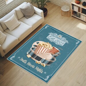 Puede incluir: Una alfombra rectangular azul con tem&aacute;tica de cine. Presenta una cubeta de palomitas de ma&iacute;z, una bebida, una bobina de pel&iacute;cula y un boleto. La alfombra tiene el texto "Smith Movie Nights" y "Est. 2023".