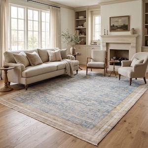 May include: A large rectangular area rug featuring a central field of light blue with intricate cream-colored floral and vine patterns. The rug has a wide, decorative border in shades of beige and tan, finished with short fringe along the edges.