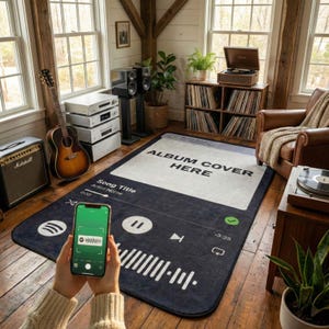 Op de afbeelding: Een groot, rechthoekig vloerkleed met een ontwerp van een muziekspelerinterface. Het vloerkleed is voornamelijk donkerblauw met witte accenten, met de tekst "ALBUM COVER HERE" en "Song Title Artist Name." Een smartphone toont een muziek-app.