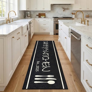 Peut inclure: Tapis de cuisine noir avec texte blanc "TAMMY'S KITCHEN" et "Est...2025". Bordure blanche et illustration de fourchette, couteau et cuillère. Posé sur un sol en bois, parfait pour une cuisine.
