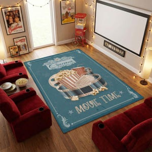 Puede incluir: Alfombra rectangular de color turquesa con un tema de cine vintage. La alfombra presenta un dise&ntilde;o de cubo de palomitas de ma&iacute;z, bebida, carrete de pel&iacute;cula y boleto. Tambi&eacute;n se muestran las palabras "Cinema" y "Movie Time". La alfombra est&aacute; rodeada de asientos rojos estilo teatro.