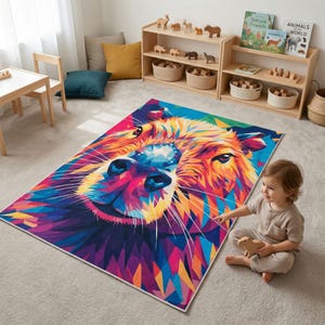 May include: A rectangular area rug featuring a vibrant, geometric design of a llama's face in shades of blue, pink, orange, and yellow. The rug is placed on a light gray carpet in a room with wooden shelves and children's books.